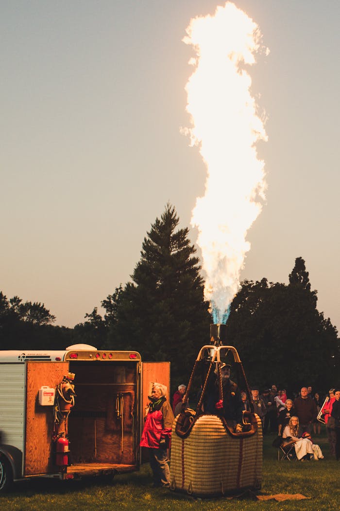 gallery-01 A group prepares a hot air balloon burner for flight at twilight, surrounded by trees and spectators.